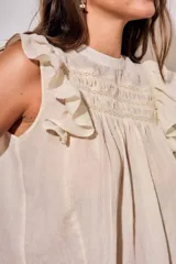 Blusa sin mangas de color crudo, con diseño oversize, cuello redondo y detalles de nido de abeja bordado en el pecho, acompañado de volados en los hombros.