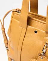 Cartera mediana de símil cuero color habano, con doble asa corta de mano y correa ajustable extraíble. Cuenta con un bolsillo frontal con broche a presión, herrajes metálicos plateados y un llavero colgante en forma de corazón.
