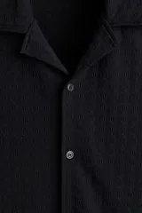 Camisa negra de manga corta con cuello resort, tapeta lisa y bajo recto con aberturas laterales. Corte holgado.