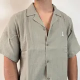 Camisa de hombre color verde oliva, de muselina con textura arrugada, manga corta, corte oversize, cuello cubano y cierre con botones. Presenta un pequeño logo bordado en el pecho.