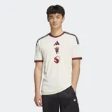 Camiseta de fútbol Adidas del Manchester United, correspondiente a la tercera equipación. Es de color blanco marfil con detalles en bordó y negro en el cuello, puños y hombros. Cuenta con el logotipo de Adidas, el escudo del club y un gráfico alineados verticalmente en el centro del pecho.