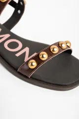 Sandalias planas de cuero marrón con tiras decoradas con tachas semiesféricas doradas, talón cerrado con tira ajustable y hebilla metálica.