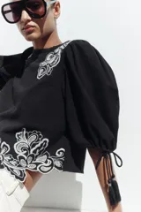 Blusa corta negra con mangas abullonadas y bordados florales en contraste de color blanco. Presenta cuello redondo con puntilla, mangas ajustables con cordones y borlas, y cierre en la espalda con abertura y botón oculto.
