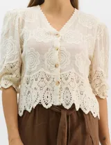 Blusa de algodón semitransparente con diseño floral bordado a tono. Presenta escote en V, cierre frontal con botones y mangas al codo ligeramente abullonadas. Incluye detalles de encaje calado estilo macramé en los puños y en el ruedo con terminaciones en ondas.