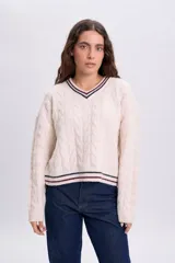 Buzo tejido de punto color crudo, con cuello redondo, puños y ruedo acanalados con rayas en contraste. Presenta un diseño trenzado en la trama y un gran número '96' aplicado en la espalda.