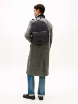 Mochila Tommy Hilfiger de diseño minimalista y silueta tipo domo en color gris oscuro. Cuenta con compartimento principal con cierre de cremallera, un bolsillo exterior frontal con solapa y cierre magnético, correas de hombro acolchadas y ajustables, y un asa superior. Presenta el logo de la marca en relieve en el frente y detalles distintivos.
