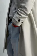 Trench coat de corte relajado, color gris claro, confeccionado en tejido técnico con mezcla de algodón. Presenta cuello solapa, manga larga con puño ajustable, bolsillos de doble vivo en cadera, bolsillo interior y abertura en el centro de la espalda. Cierre frontal con botonadura oculta.