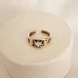 Anillo de bronce dorado con diseño abierto y una zirconia blanca engarzada en el centro. Dos pequeñas esferas de bronce decoran cada lado de la piedra.