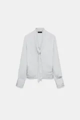 Camisa de escote en pico con lazada frontal en el mismo tejido, manga larga con puños y bajo drapeado. Presenta un acabado satinado y cierre lateral con cremallera oculta.