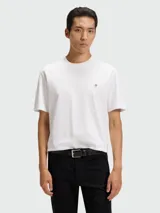 Remera de corte regular, color blanco, con cuello redondo y detalle de logo metálico circular en el pecho.