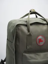 Mochila color verde militar con diseño clásico y minimalista. Cuenta con un compartimento principal con cierre de doble cremallera, un bolsillo frontal con cierre y dos asas superiores con broche. En el frente, tiene un parche circular con el texto "FORUM INDUSTRIES CO." en rojo.