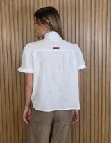 Camisa de manga corta color beige, con cuello alto con volados, botonadura frontal y mangas abullonadas con terminación fruncida.