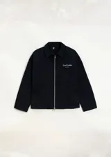 Camisa de gabardina negra de diseño unisex y corte holgado. Cuenta con un cierre frontal de doble camino, cuello camisero y detalles de estampa y bordado en el pecho con el texto 'Low Profile'.