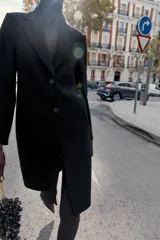 Abrigo de paño color beige con cuello solapa, manga larga con hombreras y cierre frontal con botones. Presenta bolsillos delanteros de doble vivo y abertura en la espalda.