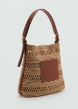 Bolso tipo hobo confeccionado en tejido de fibra natural con diseño calado. Presenta un parche central de cuero sintético, asa de hombro ajustable con hebillas metálicas y cierre magnético.