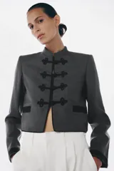 Blazer de corte cropped confeccionado en tejido de lana gris, con cuello redondo y cierre frontal mediante alamares decorativos. Presenta bolsillos delanteros con solapa y puños a contraste en color negro.