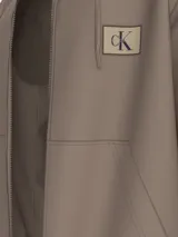 Buzo con capucha color marrón claro, con cierre frontal, bolsillos laterales y puños y dobladillo acanalados. Presenta un parche con el logo de Calvin Klein en el pecho.