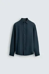 Camisa de corte relajado confeccionada en tejido fluido con mezcla de viscosa. Presenta cuello italiano, manga larga con puños abotonados y cierre frontal con botonadura, destacando por el detalle de jaretas verticales a ambos lados de la tapeta.