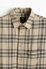 Camisa de manga larga con cuello inglés, tapeta lisa y bolsillo superior de parche. Presenta un patrón de cuadros en tonos beige claro y negro.