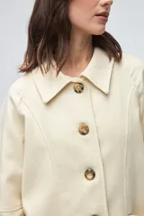 Chaquetón de lana fina sin teñir, con cuello camisero, cierre frontal de botones y bolsillos delanteros. Diseño de corte recto y estructura clásica.