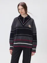 Pullover de textura tipo terciopelo con cuello alto y cierre frontal parcial. Presenta un diseño a rayas horizontales en tonos grises y negros, con detalles en rojo y blanco en el cuello, puños y ruedo. Incluye un aplique bordado con la letra M en el pecho y puños acanalados.