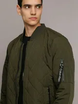 Campera bomber Calvin Klein color verde militar con diseño acolchado en forma de rombos. Tiene cierre frontal con cremallera, cuello, puños y dobladillo acanalados, bolsillos laterales y un bolsillo con cremallera en la manga izquierda con el logo de la marca.