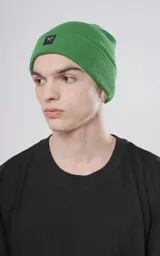 Gorro de tejido de punto color verde, de estilo beanie con borde doblado y un pequeño parche negro con un logo blanco de tijeras cruzadas.