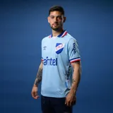 Camiseta de fútbol Umbro del Club Nacional de Football, modelo Away 3. Diseño en color celeste con cuello polo tricolor y detalles en azul, blanco y rojo en las mangas. Incluye logo de Umbro, escudo del club y sponsors estampados en el frente y laterales.