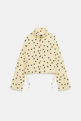 Chaqueta de cuello subido y manga larga con estampado de lunares negros sobre base beige. Cuenta con bolsillos de vivo en los laterales, detalles de frunces ajustables con cordones en el delantero y cierre frontal con botones.