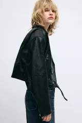 Cazadora tipo biker de corte corto y diseño holgado con hombreras. Presenta cuello con solapas, cierre frontal cruzado con cremallera, bolsillos laterales con cremallera y un bolsillo pequeño con solapa y botón a presión. Incluye un cinturón ajustable en el bajo y cremalleras en los puños.