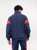 Buzo deportivo de nylon con medio cierre frontal y cuello alto. Presenta un diseño de bloques de color en azul, blanco y bordó, con el escudo del Club Nacional de Football y el logo de Umbro en el pecho.