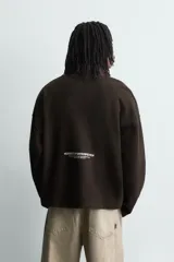 Sudadera de corte boxy fit en tejido de algodón con textura acanalada, color marrón oscuro. Presenta cuello subido con cierre frontal de cremallera y estampado frontal con texto en contraste.