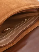 Cartera bandolera de gamuza marrón claro con solapa frontal, detalles de cuero liso más oscuro en los bordes y en el cierre de hebilla metálica dorada. Incluye correa ajustable de hombro.