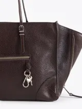 Cartera tote bag pequeña realizada en cuero vacuno marrón, con textura granulada, doble asa de hombro y bolsillos laterales con cierre. Presenta además un bolsillo frontal con cierre y detalle de tiracierres y llavero metálico colgante.