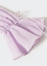 Vestido largo de tejido fluido y semitransparente color lila, con cuello en pico, mangas largas con volantes y cierre frontal con lazo. Presenta volantes decorativos a lo largo de la abertura frontal y fruncido elástico en la cintura.