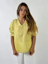 Blusa de lino color amarillo con escote en V y mangas cortas abullonadas con terminación elástica y pequeños volados. Presenta un corte holgado y textura característica del tejido de lino.