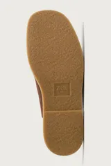 Zueco de piel con acabado serraje en color marrón, diseño sin talón, puntera redonda y detalle de costura tipo mocasín en el empeine.