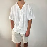 Camisa de tejido calado con textura, color blanco, de corte oversize, manga corta y cuello solapa con cierre frontal de botones.