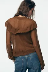 Chaqueta de punto pointelle color tostado, con cuello subido y manga larga. Presenta detalles de volantes en el pecho y puños, y cierre frontal con botones.