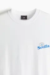 Remera de algodón blanca con cuello redondo acanalado y hombros caídos. Presenta un pequeño estampado gráfico en azul en el pecho con la palabra Sevilla.