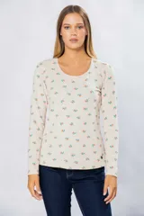 Remera de manga larga color crudo con estampado de flores pequeñas en tonos naranja y verde. Tiene cuello redondo y pin metálico con logo personalizado en el ruedo.