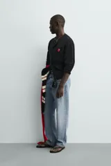 Cárdigan de punto negro, corte relajado, con cuello redondo, manga larga y cierre frontal de botones oscuros. Presenta un parche bordado en forma de dado rojo con corazones blancos en el lado izquierdo del pecho.