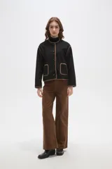 Chaqueta negra de poliéster con cuello a la base, cierre frontal con botones y dos bolsillos delanteros. Presenta detalles de bordado decorativo en color marrón en los bordes, cuello, puños y bolsillos.