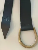Cinto de cuero flexible diseñado para anudar, con terminación en punta y detalle de marca grabada en el extremo. Incluye una hebilla metálica en forma de D en tono bronce.