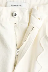 Jean de tiro regular y corte acampanado, confeccionado en denim de algodón color blanco. Presenta diseño de cinco bolsillos y detalles de costuras con ribetes bordados ondulados en el frente y los laterales.