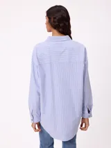 Camisa de corte oversize con diseño a rayas verticales en tonos azul y blanco. Presenta cuello clásico, cierre frontal con botones y un bolsillo en el pecho con detalle de etiqueta.