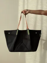 Bolso tipo tote de neopreno en color negro, con diseño estructurado y base amplia. Cuenta con correas de hombro reforzadas en tonos naranja y blanco, detalles en verde oliva y broches metálicos imantados en los laterales.