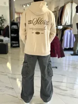 Canguro oversize de color beige con capucha y bolsillo tipo canguro al frente. En la espalda cuenta con un gran estampado tipográfico en marrón con la frase "Own Your Story", texto descriptivo y un logotipo ovalado con las letras "EST 2021".