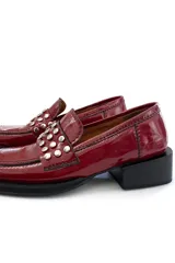 Mocasín de cuero color cereza con apliques metálicos plateados en la parte delantera, suela plana y taco cuadrado bajo color negro.