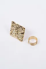 Set de dos anillos de metal dorado. Incluye un anillo con diseño de placa cuadrada de superficie texturizada y un anillo abierto de diseño minimalista con tres bandas finas paralelas.